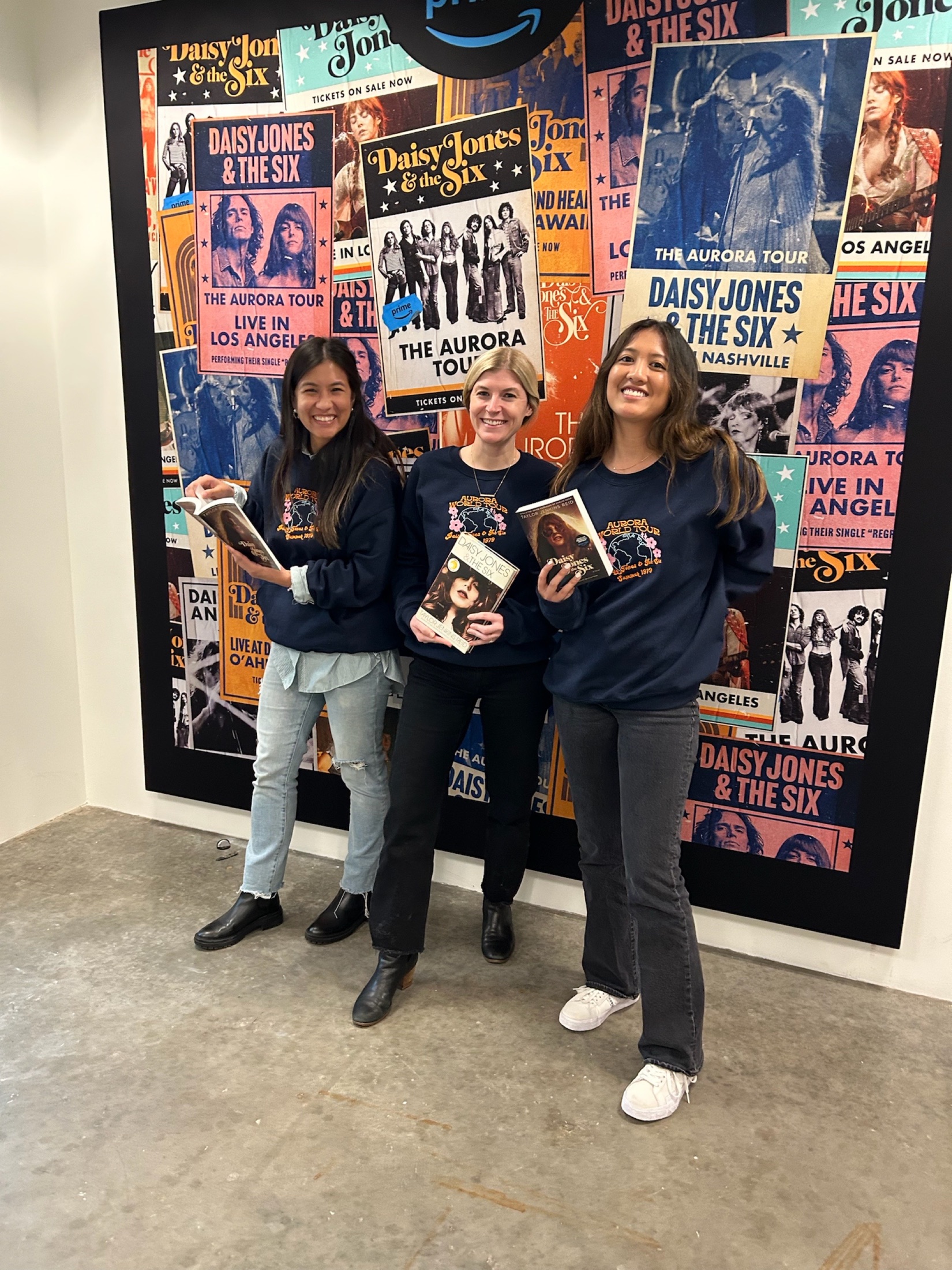 Three team members stand in front of a Daisy Jones campaign poster wall holding books.