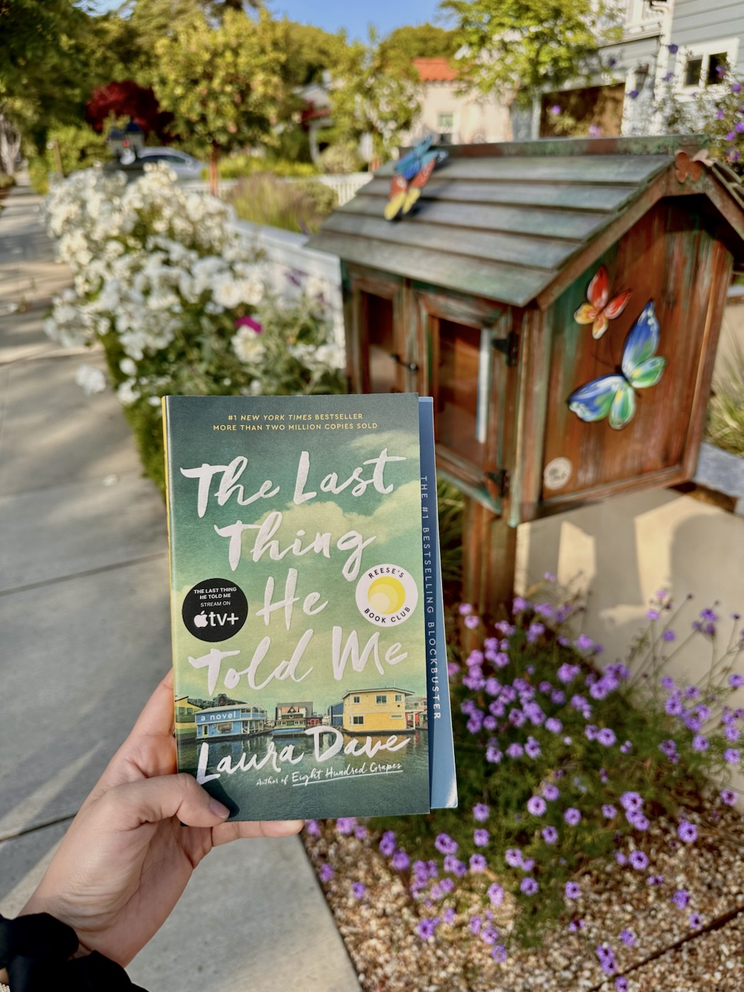 A hand holds The Last Thing He Told Me beside a neighborhood Little Free Library.