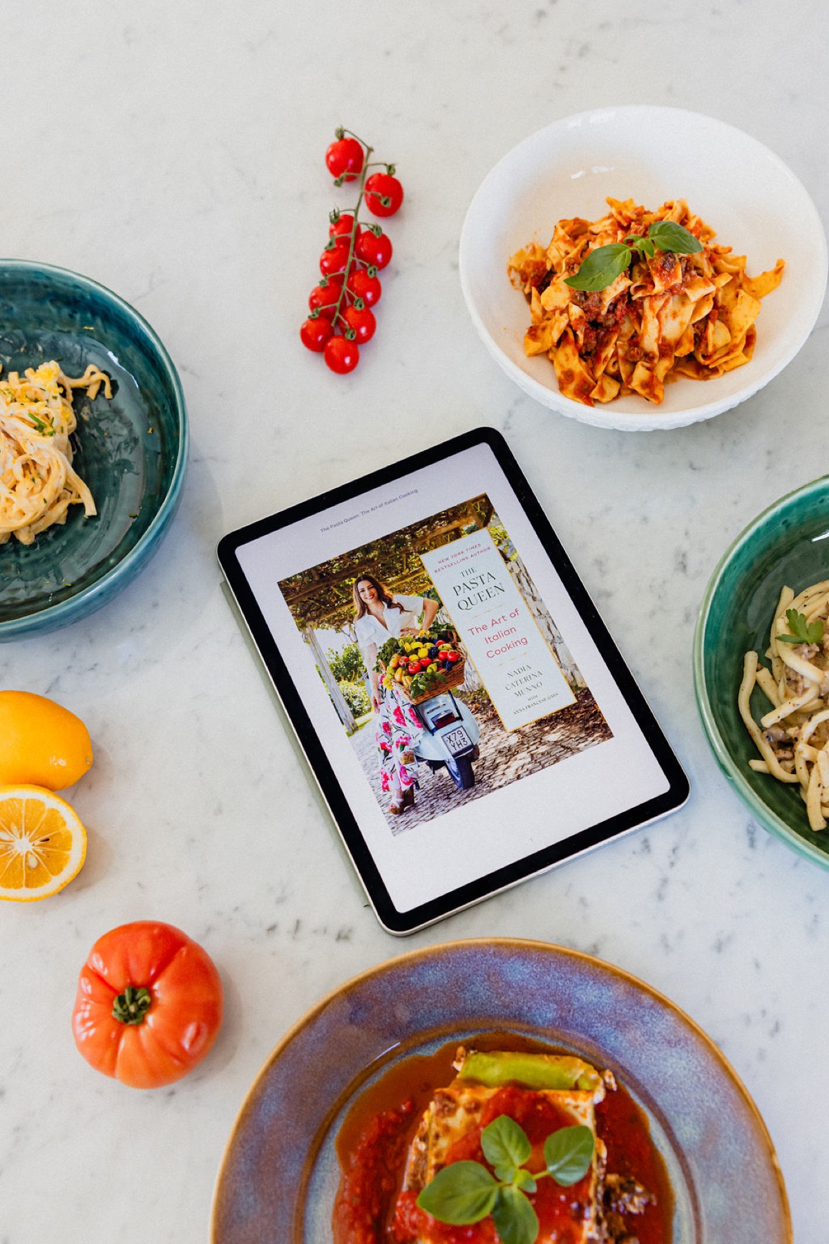 An Apple Books collection displayed on a tablet surrounded by pasta dishes.