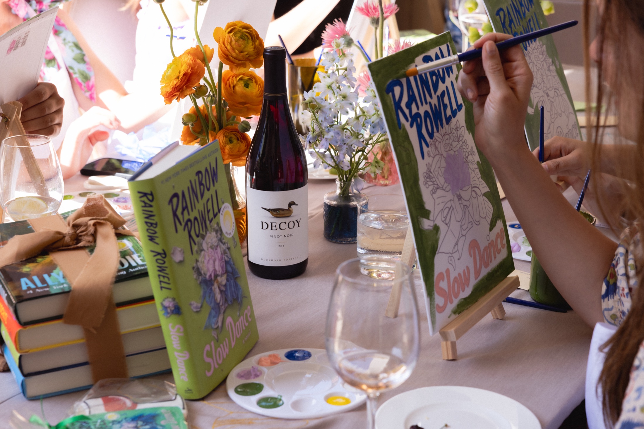 A guest painting the Slow Dance cover beside books, flowers, and Decoy wine