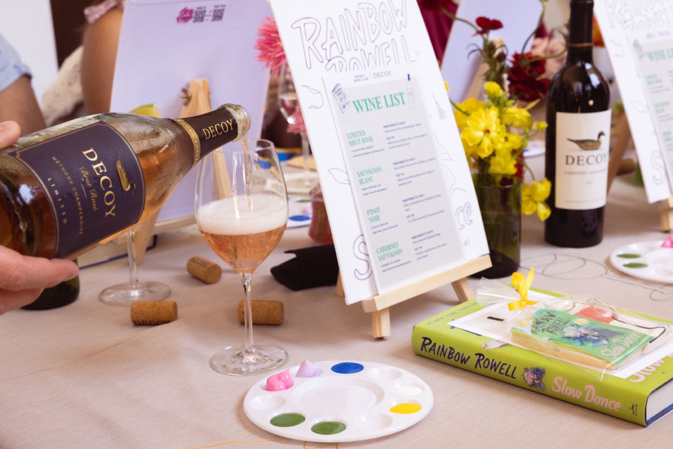 Decoy wine being poured beside the event wine list and painting supplies